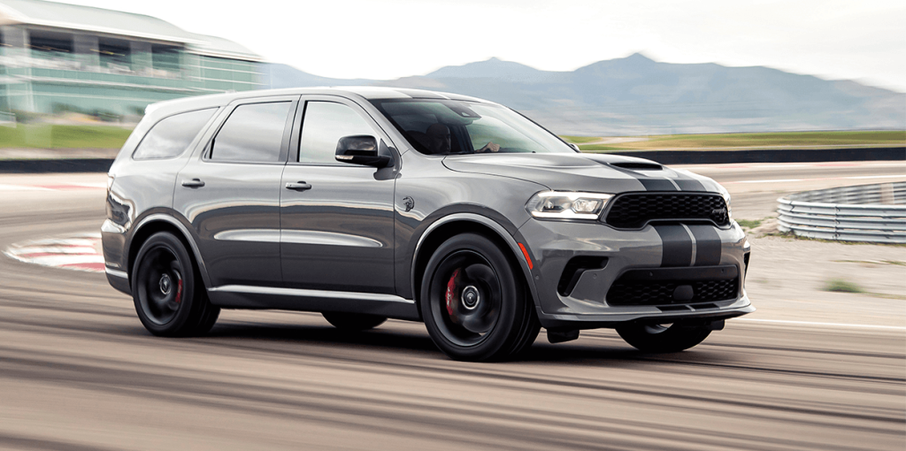 A silver 2023 Dodge Durango speeding on a racetrack with mountains and a modern building in the background.